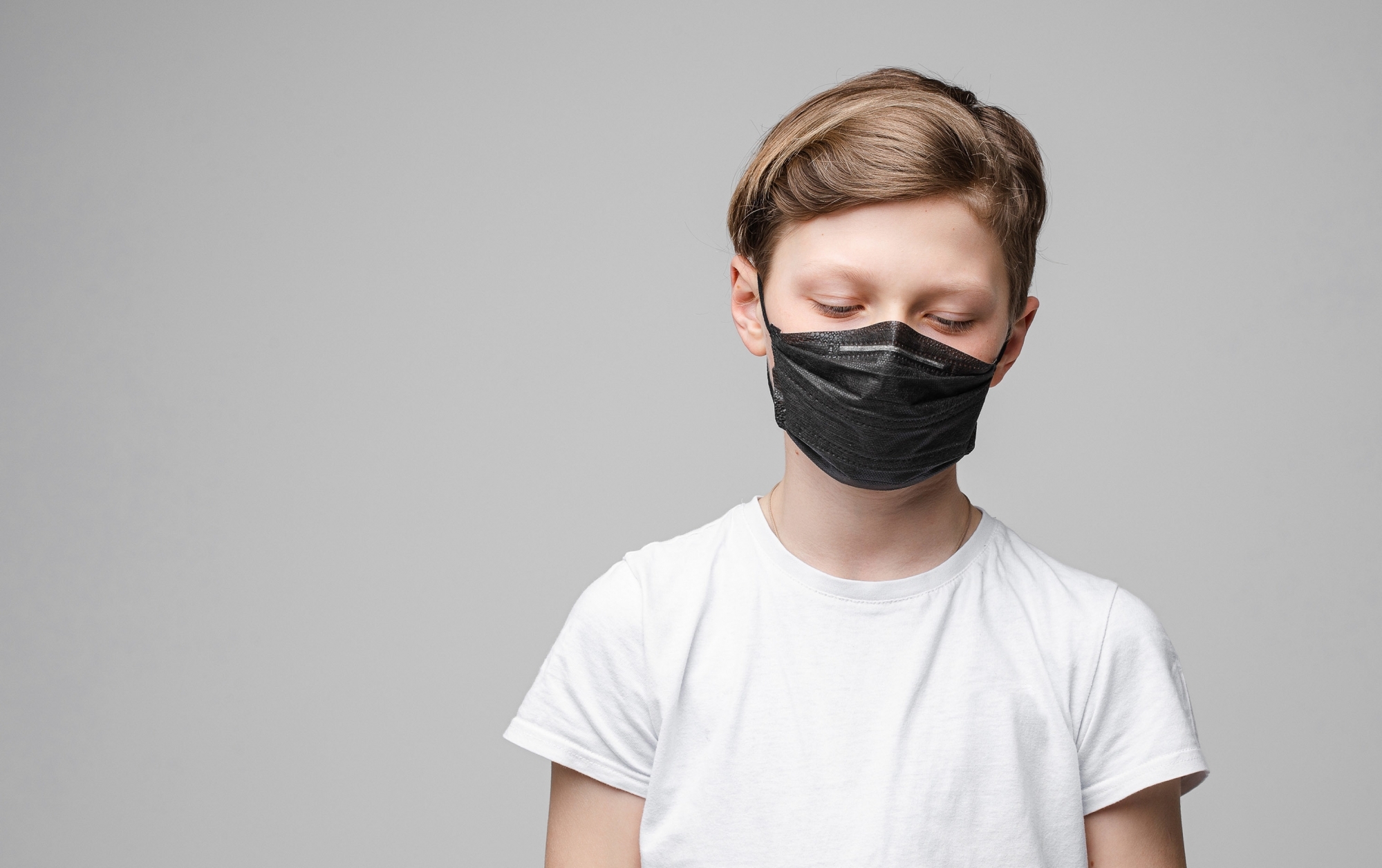 Young beautiful caucasian teenager in white t-shirt, black jeans stands with black medical mask looks down
