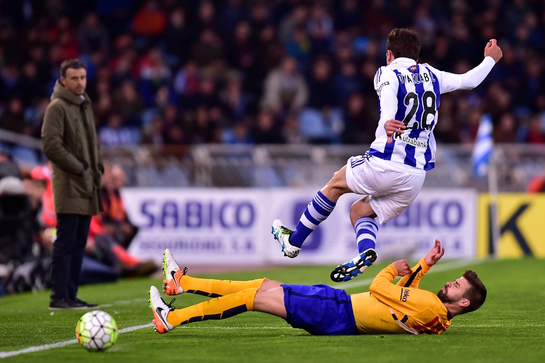 Barcelona's Gerard Pique, on the ground, duels for the ball with Real Sociedad's Mikel Oiarzabal, as head coach Luis Enrique, left, watches the action during their Spanish La Liga soccer match, at Anoeta stadium in San Sebastian, northern Spain, Saturday, April 9, 2016. (AP Photo/Alvaro Barrientos) Spain Soccer La Liga