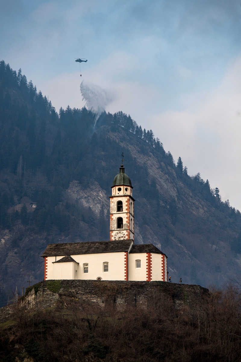 A Swiss army Superpuma Helicopter flies near the church of Soazza to discharge water over the forest fires near Mesocco in Southern Switzerland, Thursday, December 29, 2016. (KEYSTONE/Ti-Press/Gabriele Putzu) SWITZERLAND FOREST FIRE