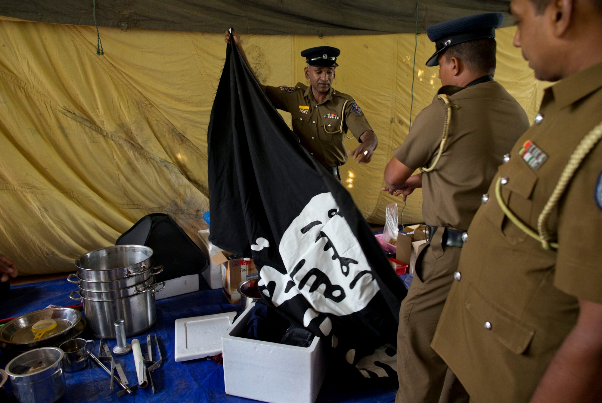 Police officers display a flag in Arabic that reads: "There is no god, but Allah" and "Of Allah is the Prophet, Muhammad" in Ampara, Sri Lanka, Sunday, April 28, 2019. Police in Ampara showed The Associated Press on Sunday the explosives, chemicals and Islamic State flag they recovered from the site of one security force raid in the region as Sri Lanka's Catholics celebrated at televised Mass in the safety of their homes. (AP Photo/Gemunu Amarasinghe) Sri Lanka Blasts